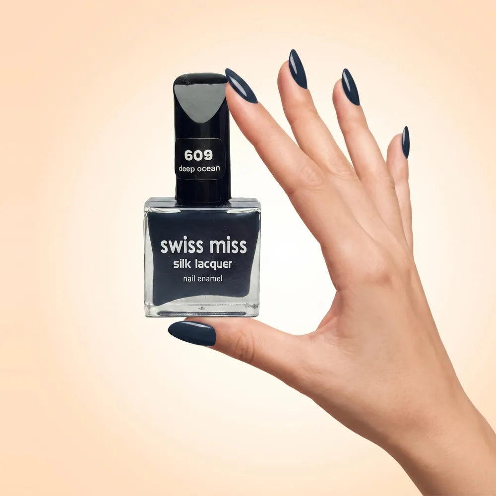 Swiss Miss Silk Lacquer Nail Enamel in shade Deep Ocean 609 held by hand showing clear glass square bottle with dark ocean navy blue polish and matching manicured nails.