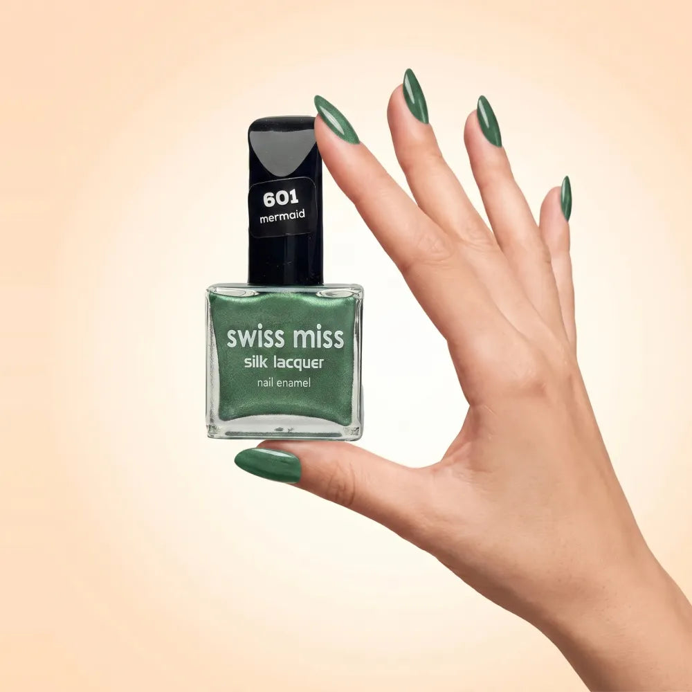 Swiss Miss Silk Lacquer Nail Enamel in shade Mermaid 601 held by hand showing clear glass square bottle with metallic emerald green polish and matching manicured nails.