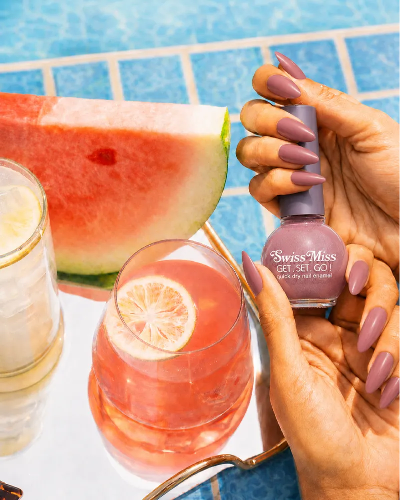 Hand holding Swiss Miss Copper Kettle 823 Get Set Go quick dry nail enamel bottle displaying metallic dusty rose copper-brown polish applied on long nails.
