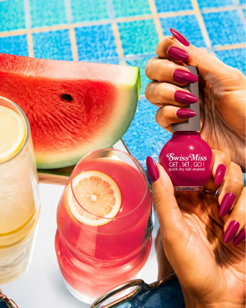 Hand holding Swiss Miss F-410 Get Set Go quick dry nail enamel bottle displaying vibrant berry magenta red polish applied on long nails.