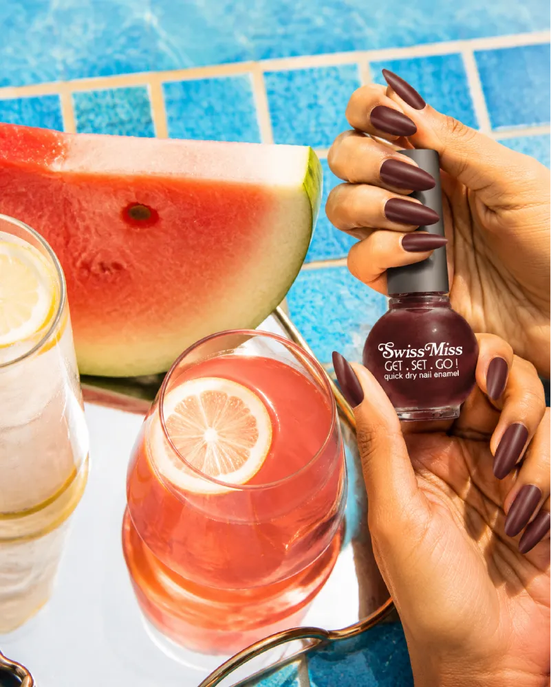 Hand holding Swiss Miss Hot Chocolate 817 Get Set Go quick dry nail enamel bottle displaying rich deep warm chocolate brown polish applied on long nails.