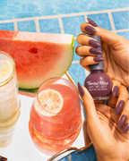 Woman holding Swiss Miss Get Set Go quick dry nail polish bottle in Moonless Nite 234 with a deep dusty mauve plum manicure by a pool.