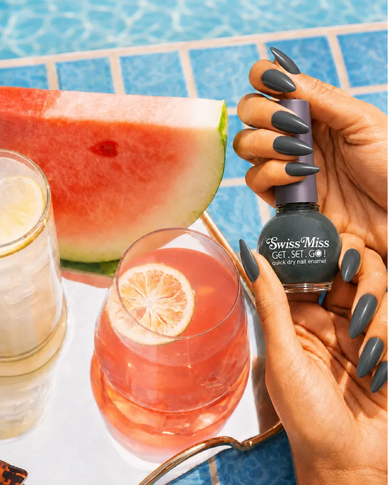 Hand holding Swiss Miss Olive Grey F-405 Get Set Go quick dry nail enamel bottle displaying cool dark olive grey polish applied on long nails.