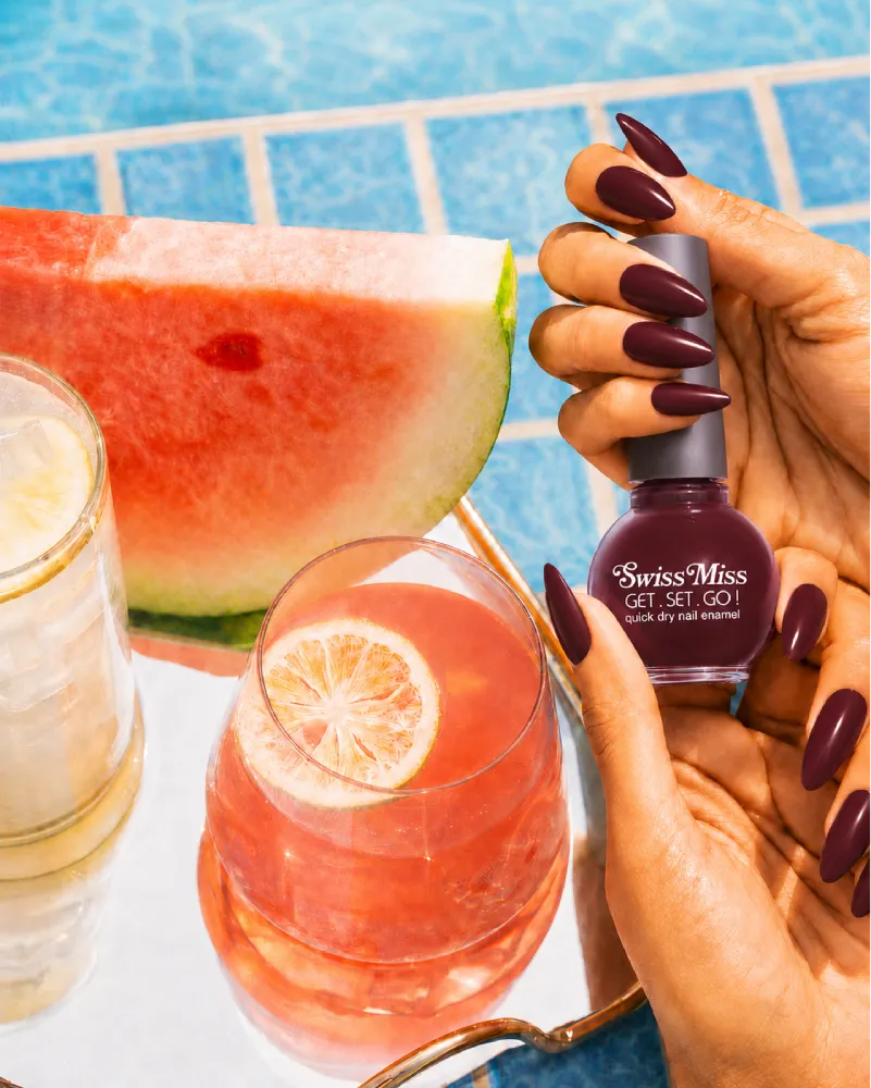 Woman holding Swiss Miss Get Set Go quick dry nail polish bottle in Ribbon Red 224 with a dark cherry red manicure by a pool.
