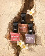 Three bottles of Swiss Miss Silk Lacquer nail enamel framed in a textured stone wall crack adorned with white plumeria flowers.
