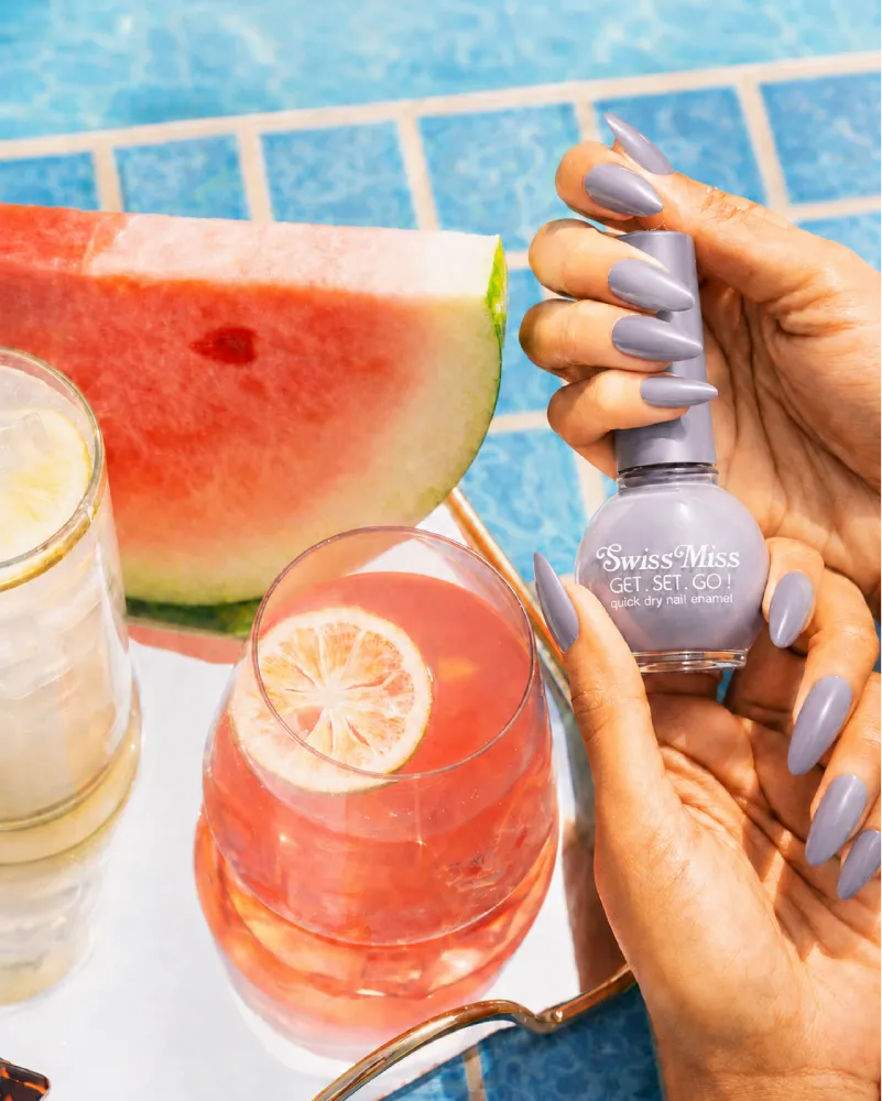 Hand holding Swiss Miss Silver Lining 812 Get Set Go quick dry nail enamel bottle displaying cool light grey silver polish applied on long nails.